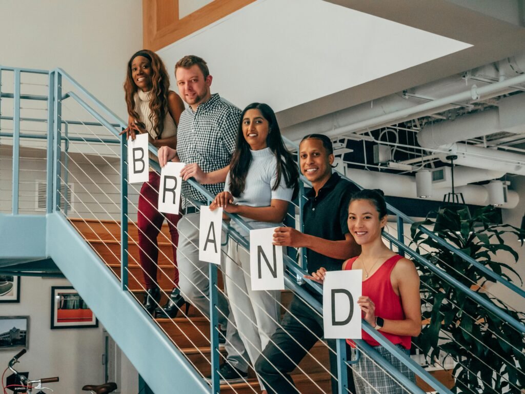 A multicultural group of colleagues smiling while holding letter signs spelling 'BRAND' on an office staircase.