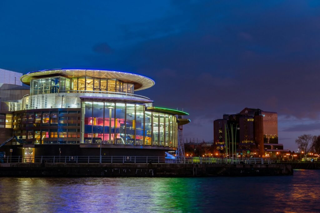 Illuminated modern building by a river in Manchester at night. Vibrant reflections enhance the structure's aesthetic.